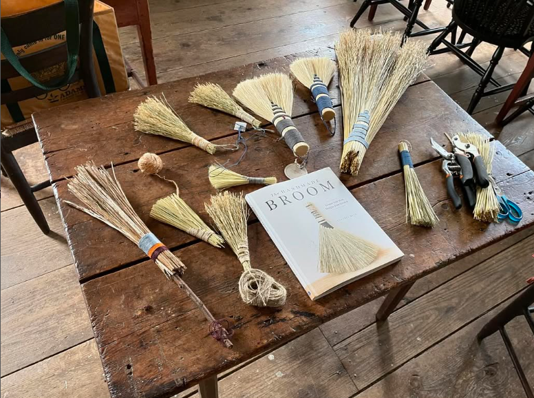 A wood table top covered in small, student made hand brooms and a book on straw broom making.